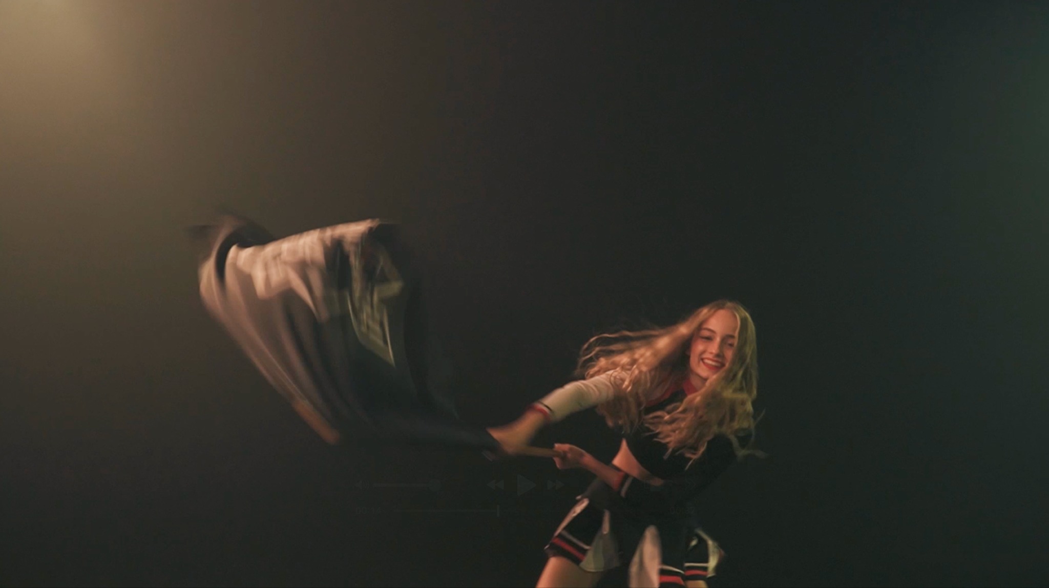 Młoda kobieta w stroju cheerleadera macha flagą na ciemnym tle, uśmiechnięta i dynamiczna.