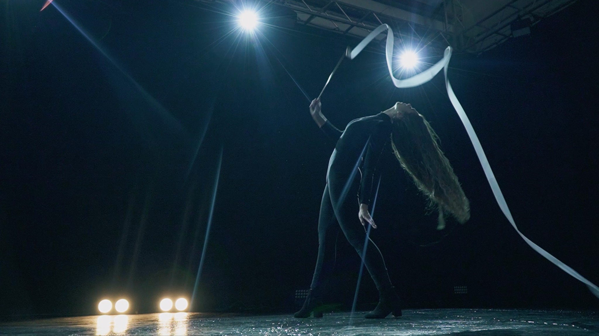 Kobieta w czarnym stroju gimnastycznym wykonuje układ z białą wstążką na scenie w blasku reflektorów.