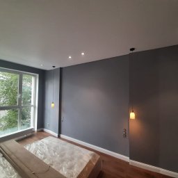 Khatai Valizada - Świeżo pomalowane szare ściany w pokoju z drewnianą podłogą, okno z widokiem na zieleń miejską, meble zabezpieczone folią, wiszące lampy z ciepłym światłem.