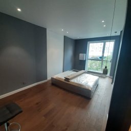 Khatai Valizada - Pokój z drewnianą podłogą, szarymi ścianami i łóżkiem w folii ochronnej, widok na okno z wiszącą lampą.