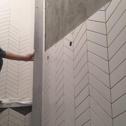 Khatai Valizada - Układanie białych, podłużnych płytek w jodełkę na ścianie łazienki; glazurnik używa poziomicy do sprawdzenia pionu; widoczne otwory na instalacje.