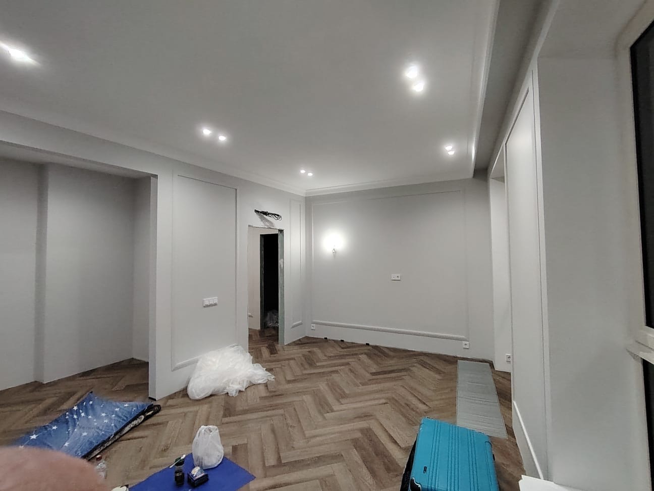 Pokój po remoncie z parkietem w jodełkę, białe ściany z dekoracyjnymi listwami, widoczne materiały budowlane i bagaż podróżny.
