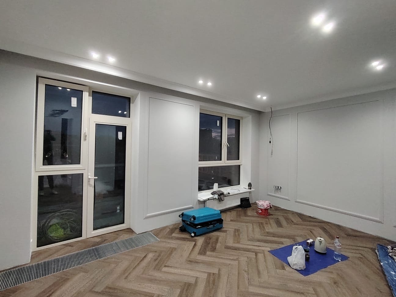 Pokój w trakcie remontu z ułożonym parkietem w jodełkę, pomalowanymi na szaro ścianami z dekoracyjnymi listwami, niebieską walizką narzędziową i akcesoriami malarskimi na podłodze.