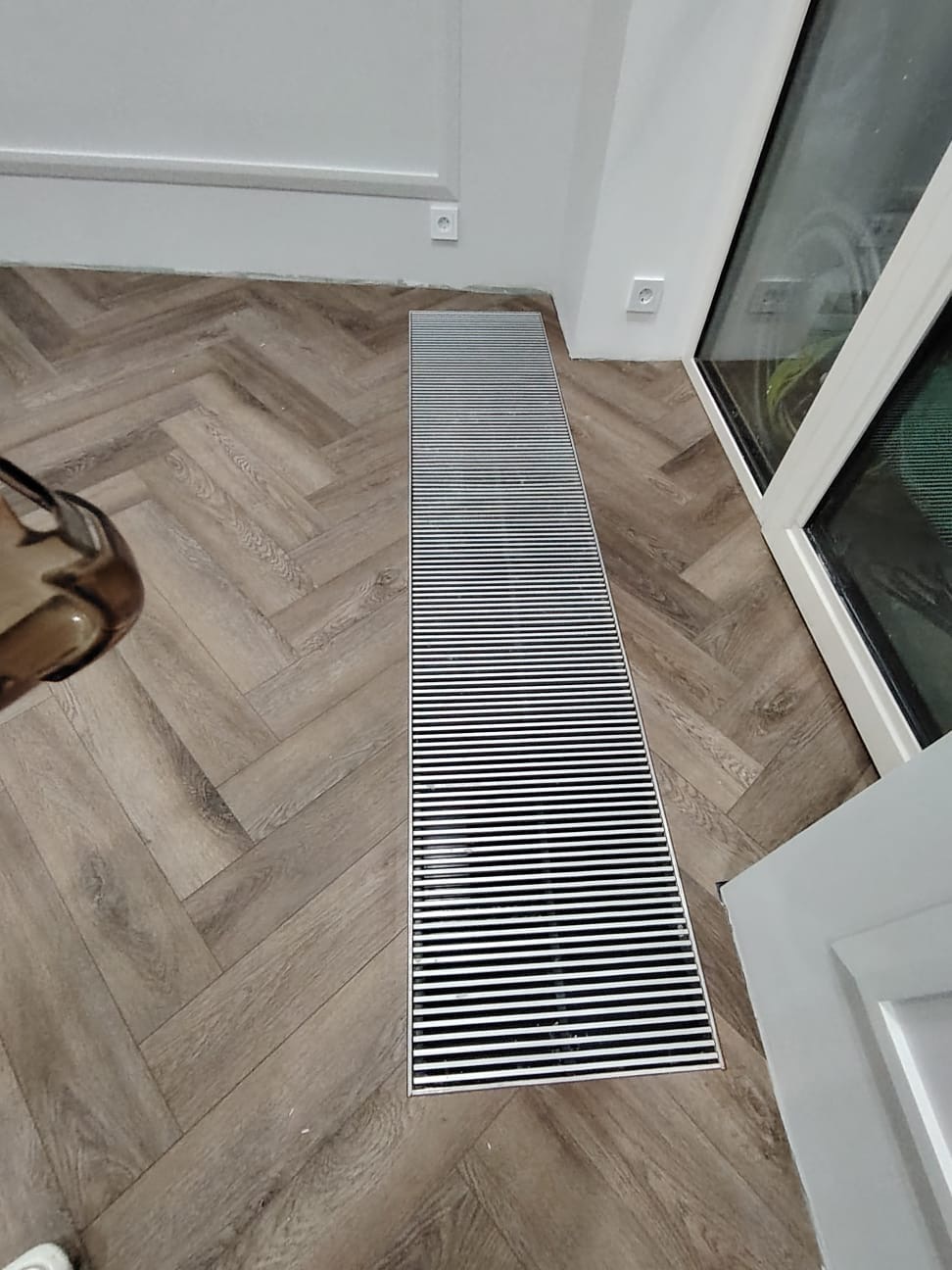 Ułożona podłoga z paneli w jodełkę, z zamontowanym liniowym odpływem podłogowym przy oknie.