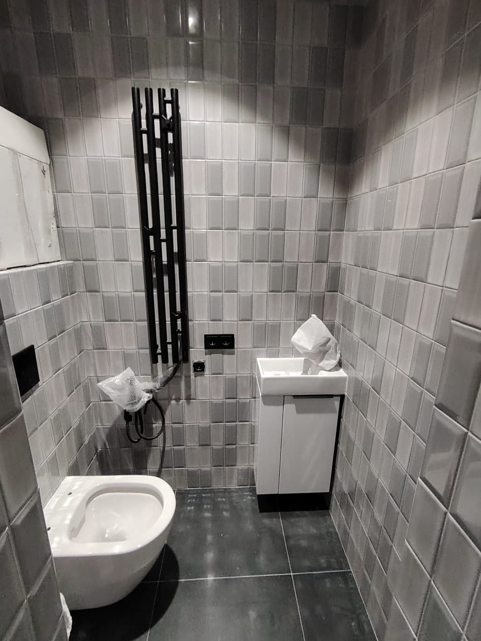 Nowoczesna łazienka w Gdańsku z szarymi płytkami ułożonymi w cegiełkę, wisząca toaleta, biała szafka z umywalką i czarny grzejnik drabinkowy. Część elementów jeszcze w folii ochronnej.