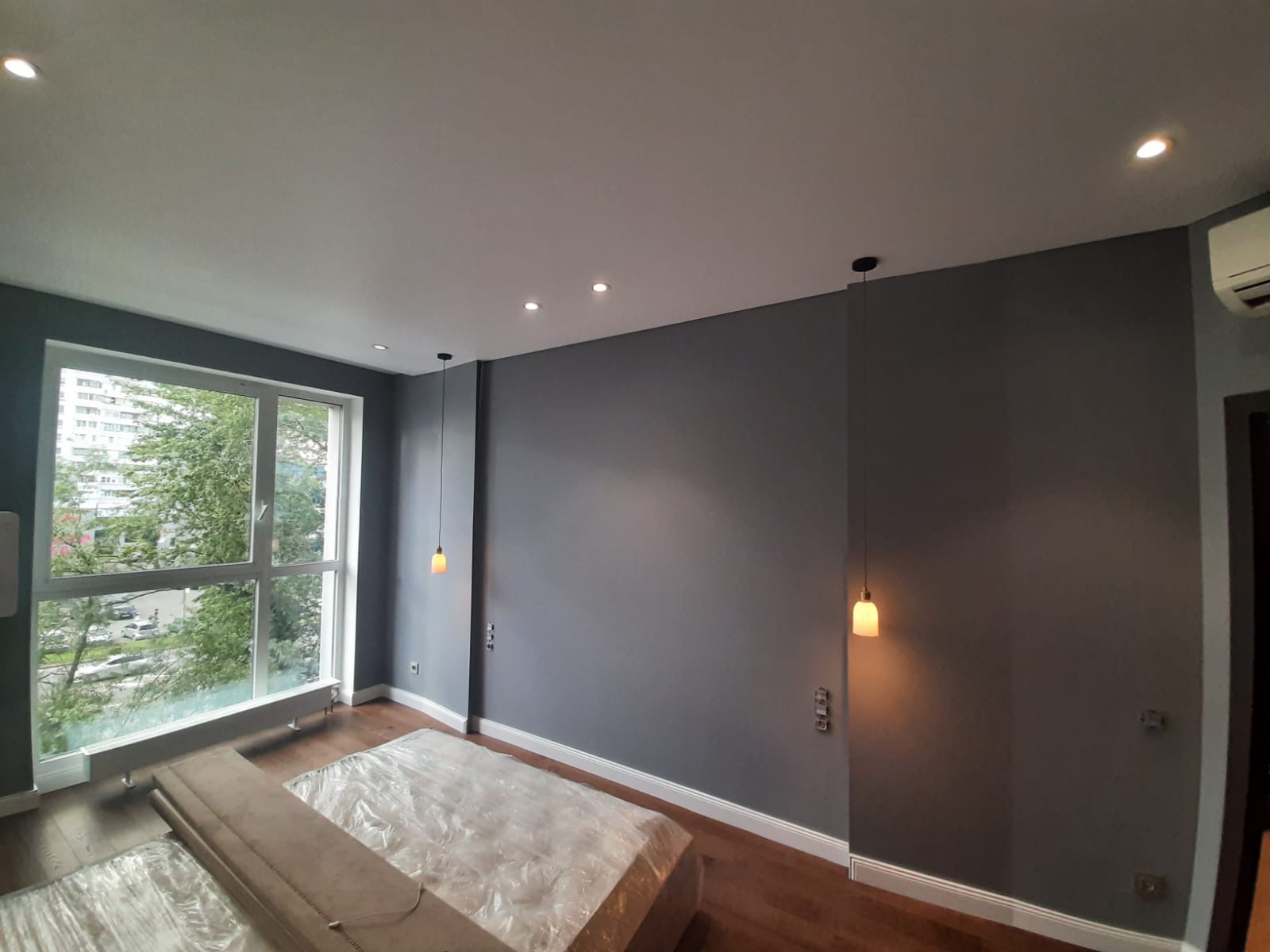 Świeżo pomalowane szare ściany w pokoju z drewnianą podłogą, okno z widokiem na zieleń miejską, meble zabezpieczone folią, wiszące lampy z ciepłym światłem.
