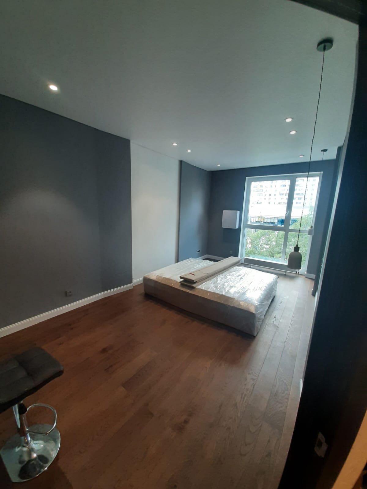 Pokój z drewnianą podłogą, szarymi ścianami i łóżkiem w folii ochronnej, widok na okno z wiszącą lampą.