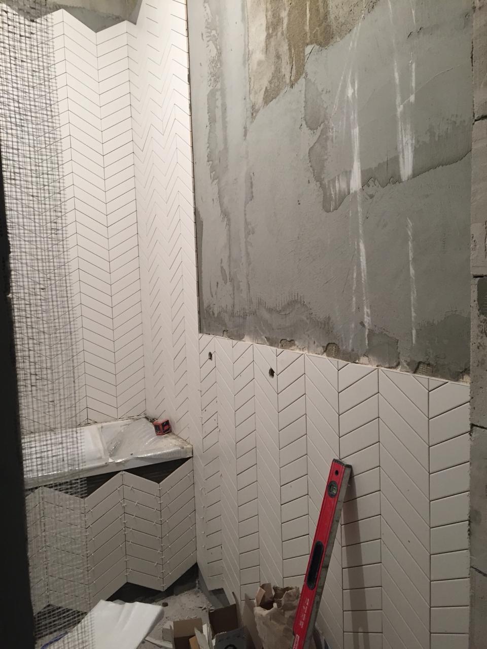 Białe, wąskie płytki ułożone w jodełkę na ścianach i obudowie wanny w trakcie prac wykończeniowych łazienki, widoczny poziomica i surowy beton na ścianie.