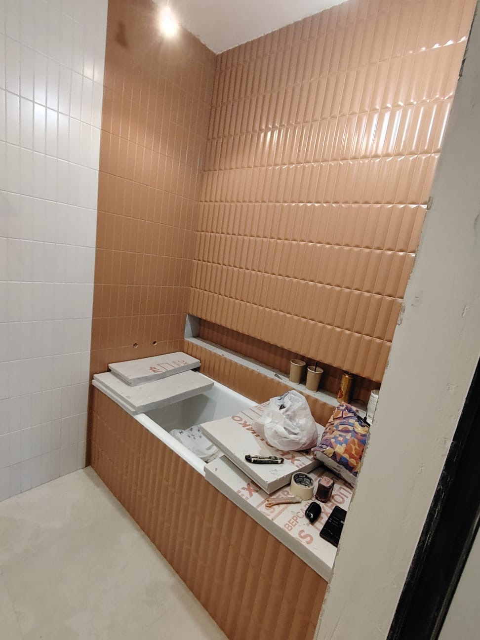 Łazienka w trakcie wykańczania: brązowe, pionowo ułożone płytki na ścianach i obudowie wanny, widoczne narzędzia i materiały budowlane, białe płytki na sąsiedniej ścianie.