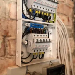 Rozdzielnica elektryczna z bezpiecznikami i przewodami w białej obudowie, zamontowana na ścianie z cegły, obok widoczne liczniki energii.