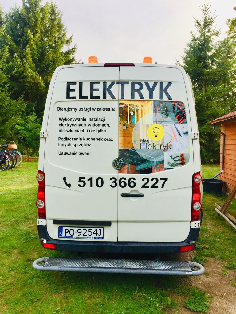 Tył białego samochodu dostawczego z napisem ELEKTRYK i numerem telefonu, prezentujący usługi elektryczne, na tle zielonych drzew i domku.