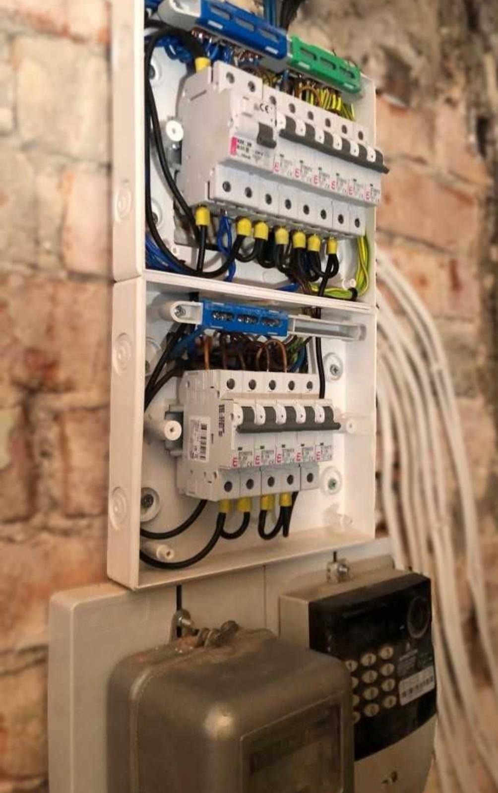 Rozdzielnica elektryczna z bezpiecznikami i przewodami w białej obudowie, zamontowana na ścianie z cegły, obok widoczne liczniki energii.