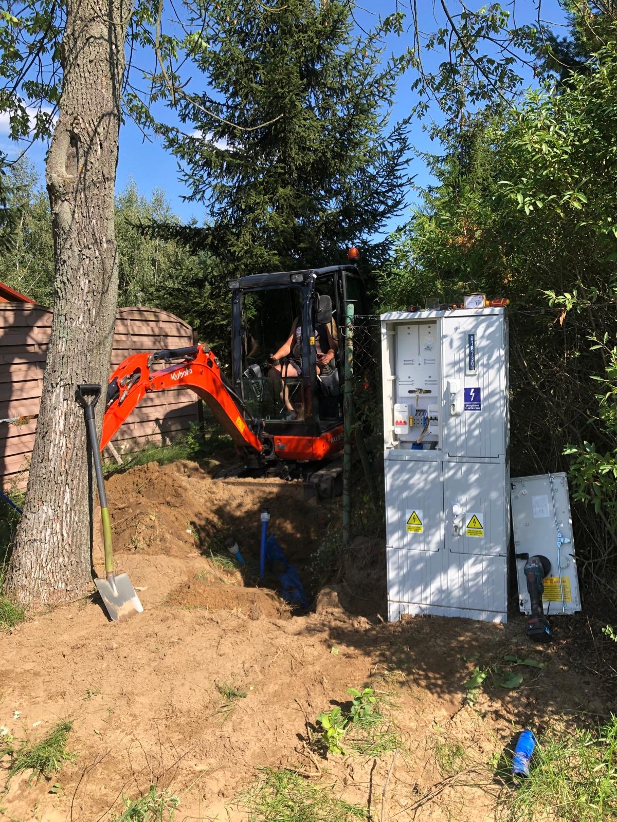 Prace ziemne minikoparką Kubota przy szafie elektrycznej z otwartymi drzwiczkami, widoczne niebieskie rury w wykopie i łopata oparta o drzewo.