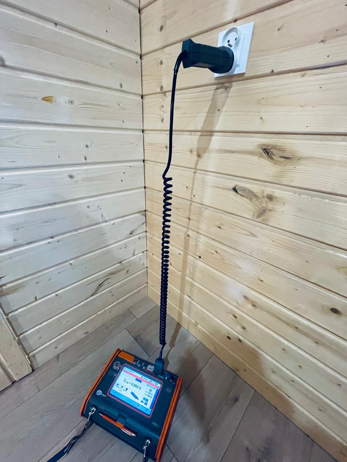 Miernik elektryczny podłączony do gniazdka w drewnianej ścianie, wyświetlający wynik pomiaru impedancji pętli zwarcia ZL=0.860 Ω.