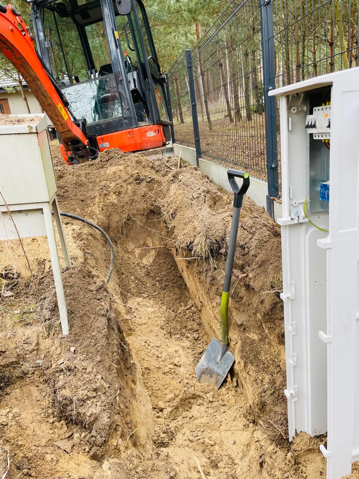 Wykopany rów w ziemi z kablem, obok stoi otwarta skrzynka elektryczna z zamontowanymi bezpiecznikami i wyłącznikami, a w tle widoczny jest minikoparka i ogrodzenie.