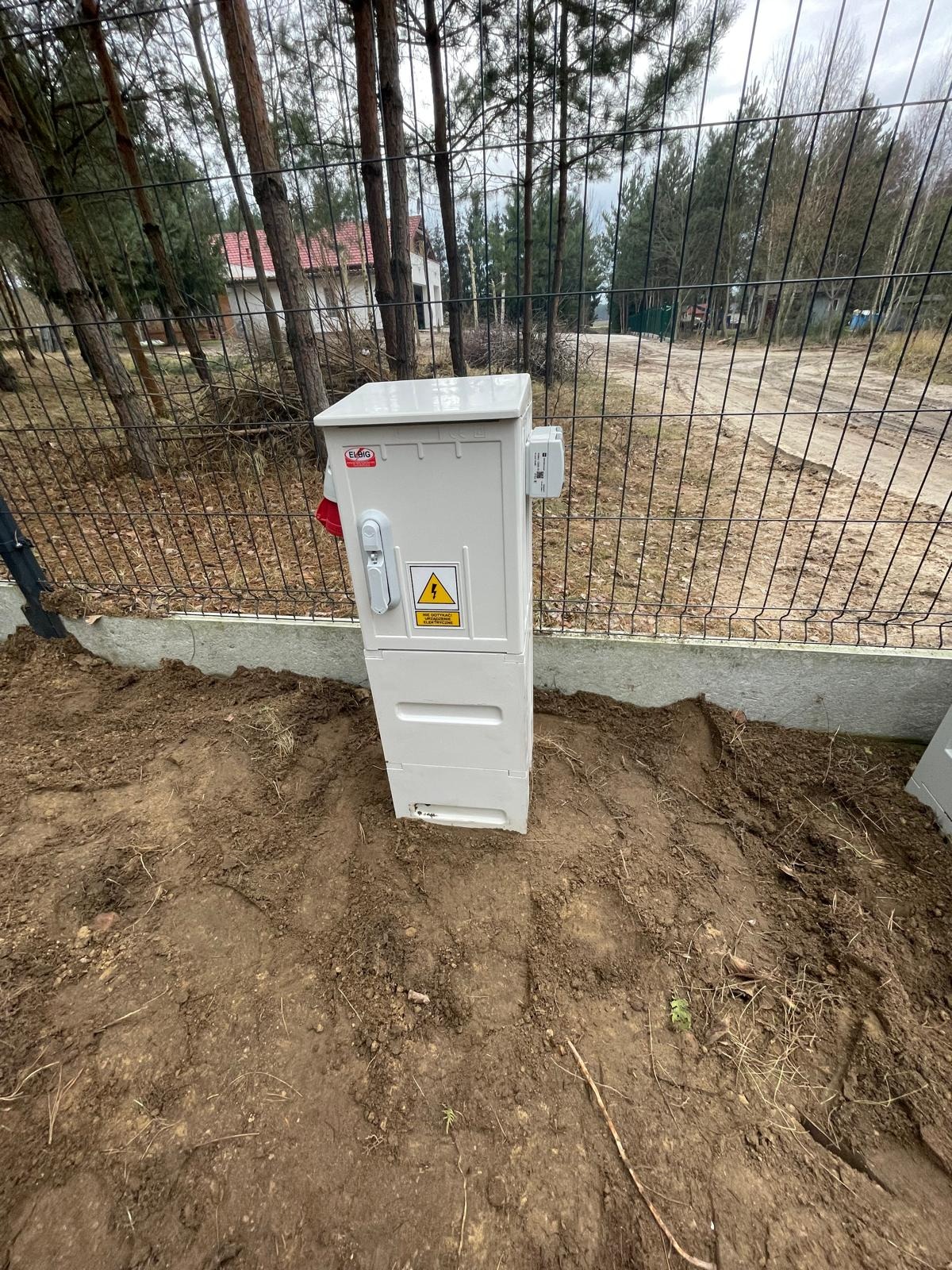 Zewnętrzna skrzynka elektryczna z zamkiem i tabliczką ostrzegawczą umieszczona na ziemi przy ogrodzeniu z siatki.