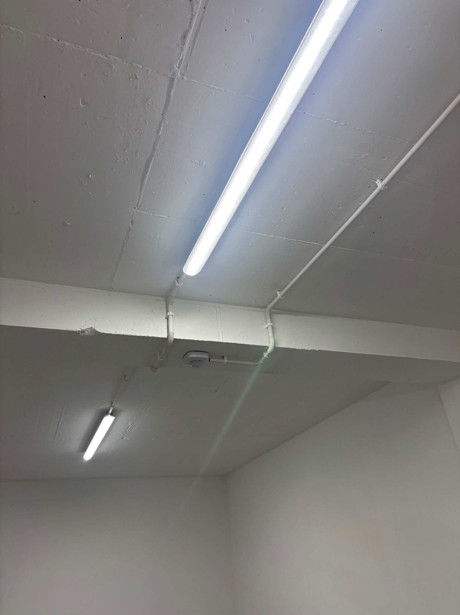 Widok sufitu z dwiema podłużnymi lampami fluorescencyjnymi, rurami instalacyjnymi i czujnikiem ruchu, wszystko w odcieniach bieli.