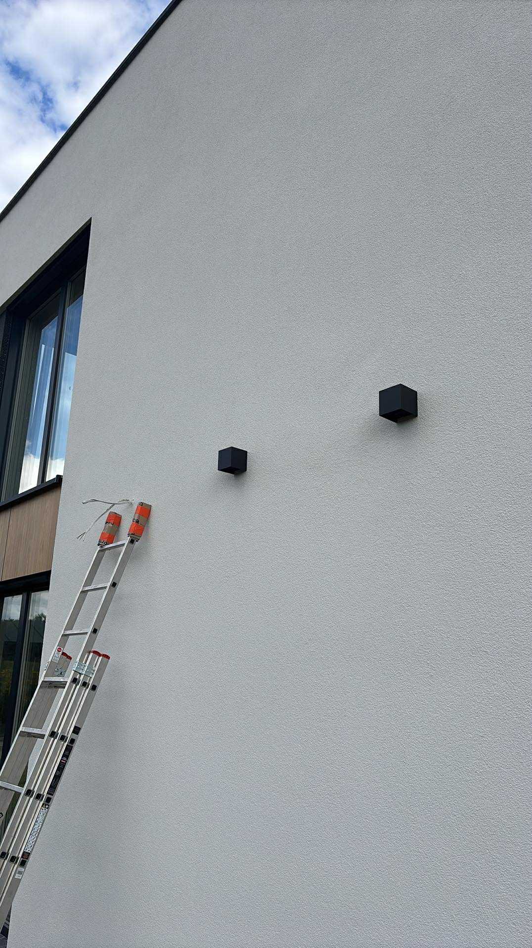 Dwa czarne, kubiczne kinkiety zamontowane na szarej, zewnętrznej ścianie budynku. Aluminiowa drabina oparta o ścianę z widocznymi przewodami.