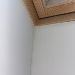 Finezja Wykończeń Krzysztof Chrobak - Biała ściana po szpachlowaniu i malowaniu, z drewnianą ramą okna dachowego. Gładka powierzchnia, widoczne łączenie ściany z oknem.