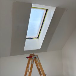 Finezja Wykończeń Krzysztof Chrobak - Pokój na poddaszu w trakcie malowania. Okno dachowe zabezpieczone folią, ściany częściowo pomalowane na biało, widoczna drabina malarska z nożem.