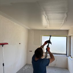 Finezja Wykończeń Krzysztof Chrobak - Praca natryskowa gładzi na suficie w nowym pomieszczeniu. Okno zabezpieczone folią, widoczne ślady farby na podłodze, przygotowanie do dalszych prac wykończeniowych.