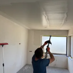 Praca natryskowa gładzi na suficie w nowym pomieszczeniu. Okno zabezpieczone folią, widoczne ślady farby na podłodze, przygotowanie do dalszych prac wykończeniowych.