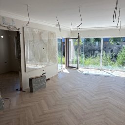 Finezja Wykończeń Krzysztof Chrobak - Jasne panele ułożone w jodełkę w przestronnym pokoju z dużymi oknami. Widoczne niedokończone prace wykończeniowe, surowe kable zwisające z sufitu i zapakowane materiały budowlane.