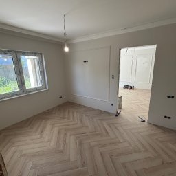 Finezja Wykończeń Krzysztof Chrobak - Jasne panele ułożone w jodełkę w pokoju z oknem i dekoracyjnymi listwami. W tle widoczne kolejne pomieszczenie z podobnym wykończeniem. Widać niedokończone prace.