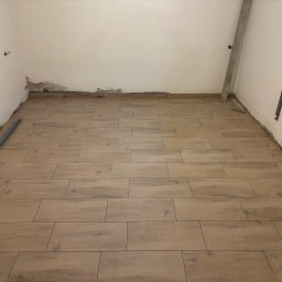 Finezja Wykończeń Krzysztof Chrobak - Ukończone układanie podłogi z drewnopodobnych płytek w jasnym pomieszczeniu. Widoczne listwy przypodłogowe i narzędzia glazurnicze. Ściany wymagają wykończenia.