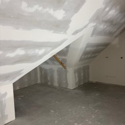 Finezja Wykończeń Krzysztof Chrobak - Poddasze w trakcie prac wykończeniowych: szpachlowane ściany i sufit z płyt gipsowo-kartonowych, widoczne łączenia i nierówności przed malowaniem. Surowy beton na podłodze.