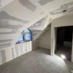 Finezja Wykończeń Krzysztof Chrobak - Poddasze w trakcie wykańczania: ściany z płyt gipsowo-kartonowych po szpachlowaniu, okno zabezpieczone folią, surowa wylewka. Widoczne otwory na oświetlenie.