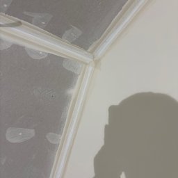 Finezja Wykończeń Krzysztof Chrobak - Narożnik sufitu po montażu listew wykończeniowych, widoczne łączenie ściany i sufitu przed malowaniem, cień osoby w prawym dolnym rogu.