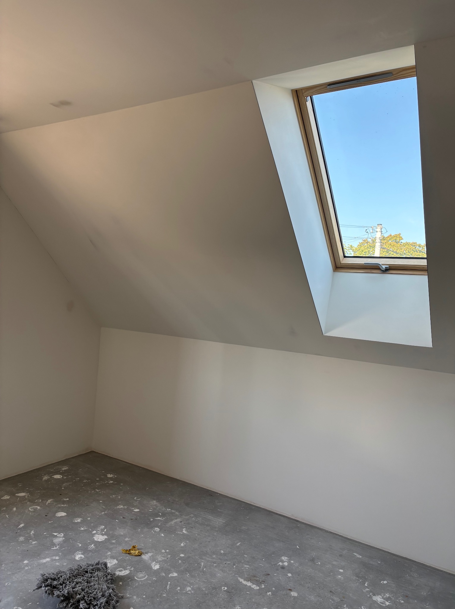 Poddasze po remoncie: białe ściany, okno dachowe z widokiem na błękitne niebo, surowa podłoga z resztkami po pracy. Minimalistyczny, jasny pokój.