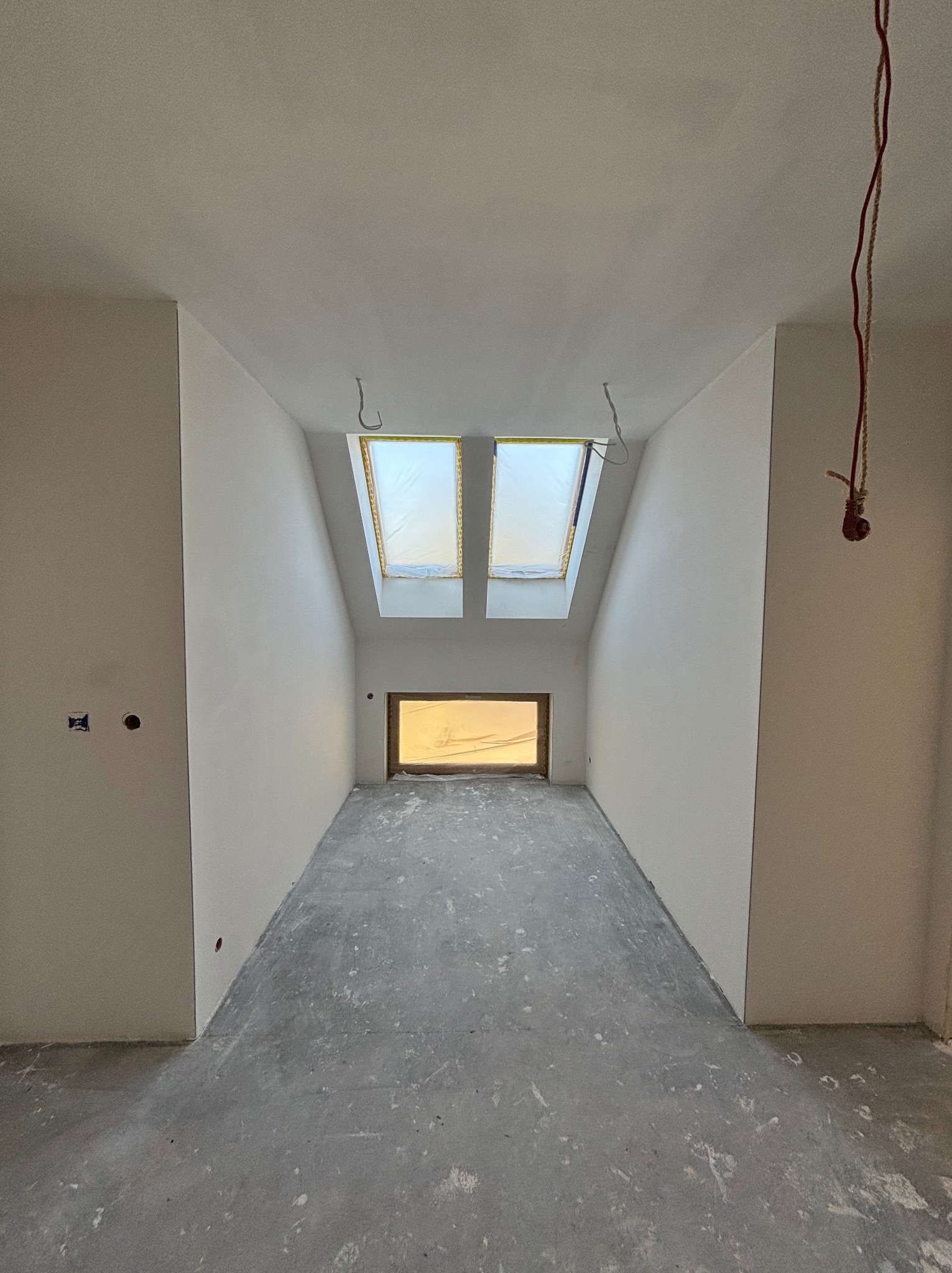 Wnętrze poddasza po gładziowaniu ścian i sufitu, z oknami dachowymi zabezpieczonymi folią i surową podłogą. Widoczne kable i puszki elektryczne.