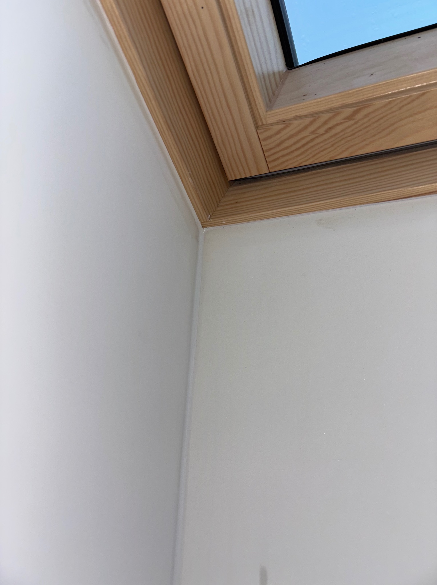 Biała ściana po szpachlowaniu i malowaniu, z drewnianą ramą okna dachowego. Gładka powierzchnia, widoczne łączenie ściany z oknem.