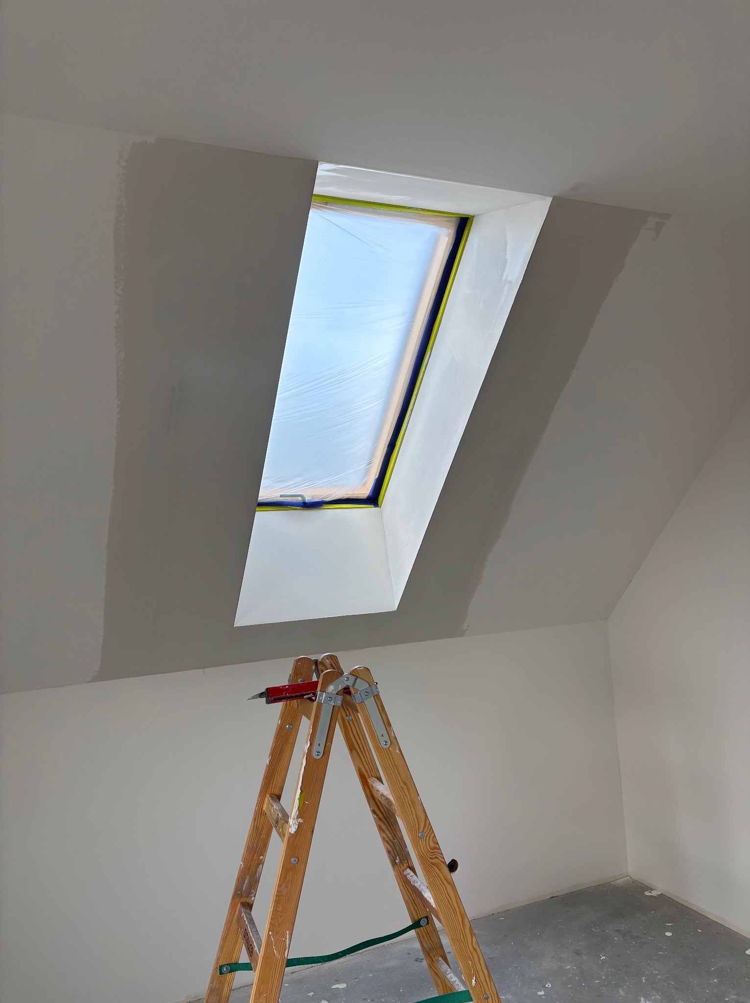 Pokój na poddaszu w trakcie malowania. Okno dachowe zabezpieczone folią, ściany częściowo pomalowane na biało, widoczna drabina malarska z nożem.