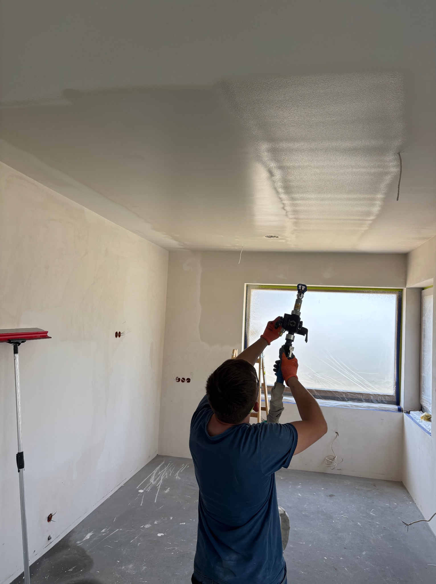 Praca natryskowa gładzi na suficie w nowym pomieszczeniu. Okno zabezpieczone folią, widoczne ślady farby na podłodze, przygotowanie do dalszych prac wykończeniowych.