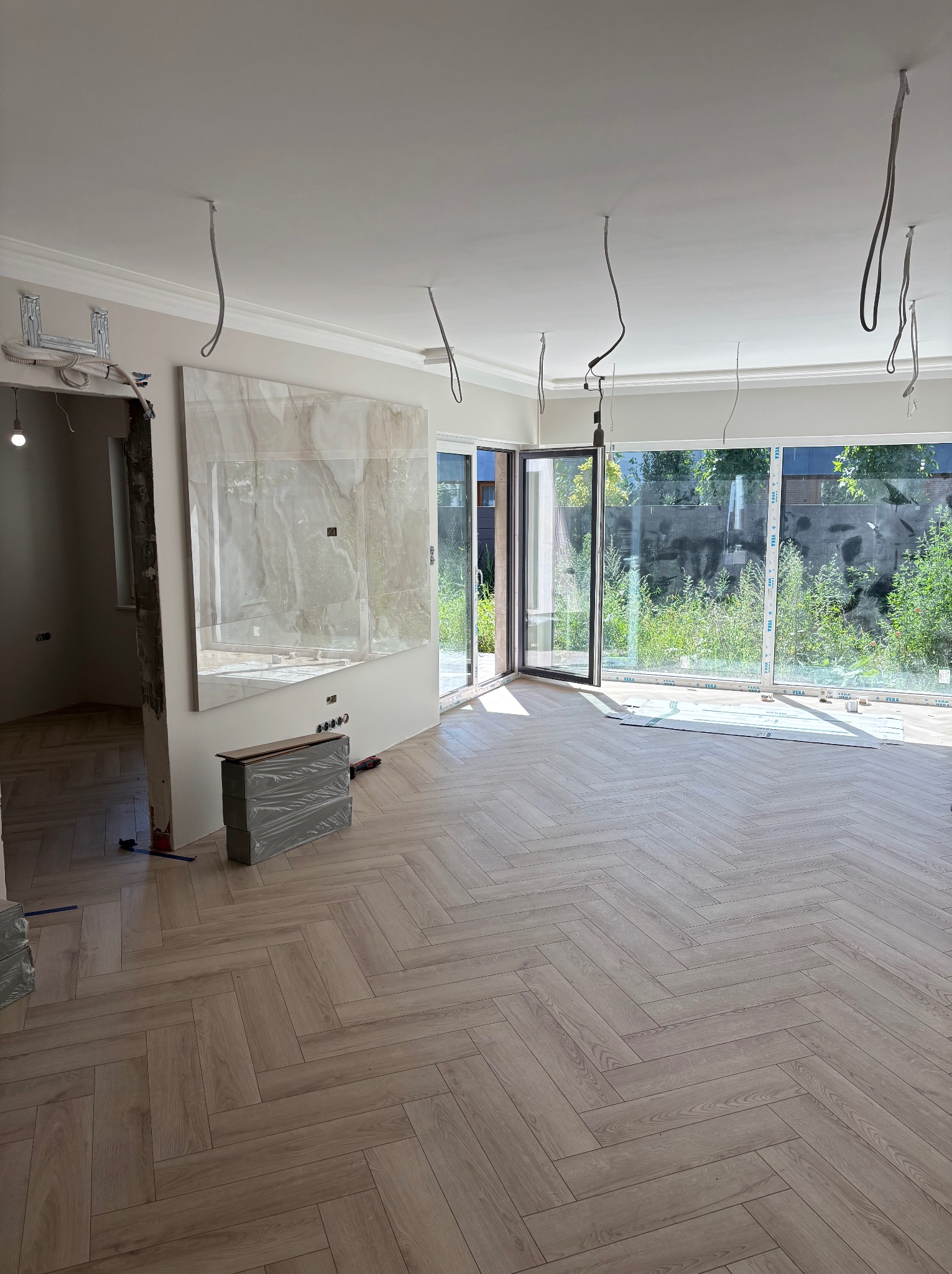 Jasne panele ułożone w jodełkę w przestronnym pokoju z dużymi oknami. Widoczne niedokończone prace wykończeniowe, surowe kable zwisające z sufitu i zapakowane materiały budowlane.