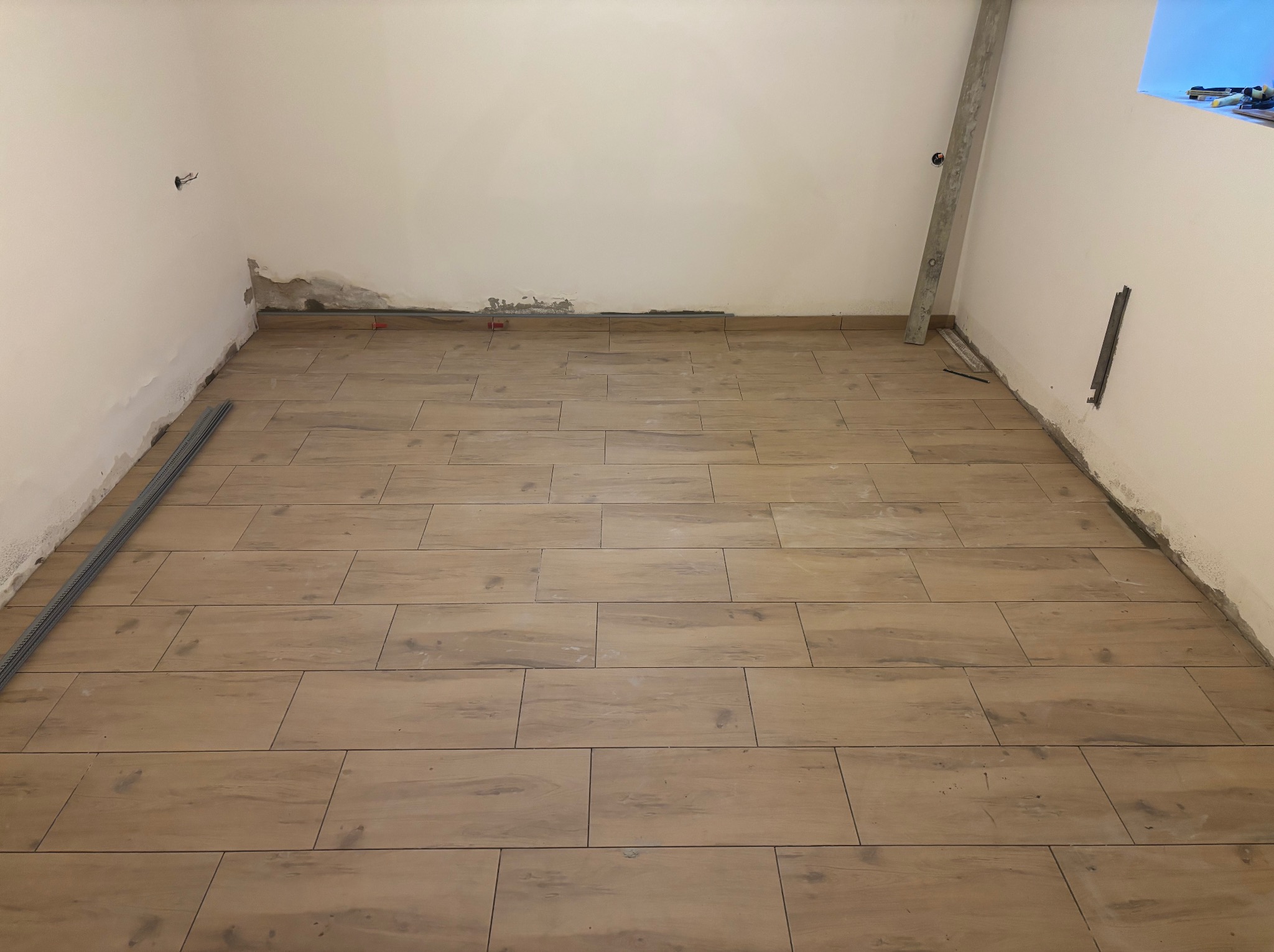 Ukończone układanie podłogi z drewnopodobnych płytek w jasnym pomieszczeniu. Widoczne listwy przypodłogowe i narzędzia glazurnicze. Ściany wymagają wykończenia.