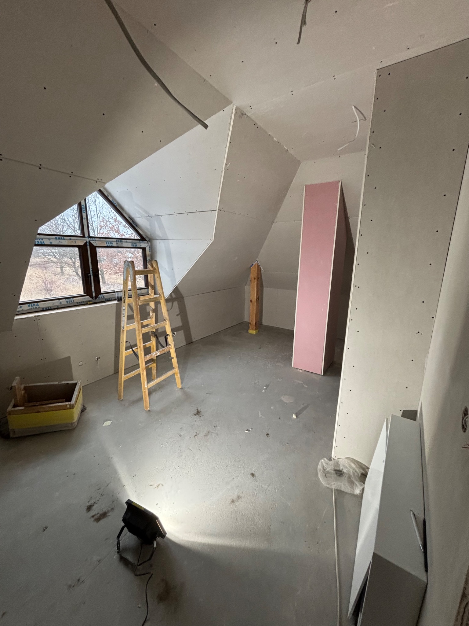 Poddasze w trakcie adaptacji: ściany obłożone płytami gipsowymi, drewniana drabina przy oknie, lampa budowlana na podłodze. Widoczne kable i elementy konstrukcyjne.