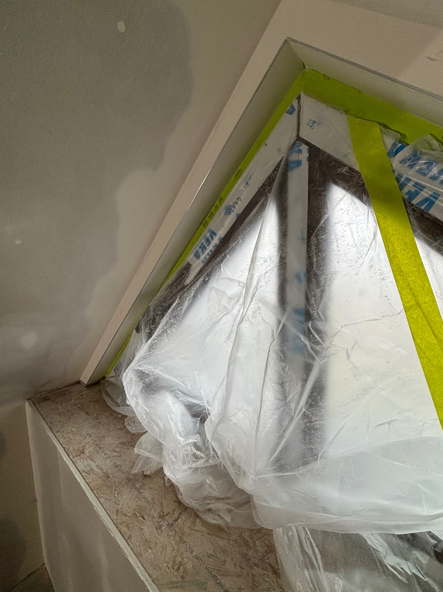 Okno zabezpieczone folią i taśmą malarską podczas prac wykończeniowych w pomieszczeniu ze ścianami z płyt kartonowo-gipsowych. Widoczny fragment sufitu.