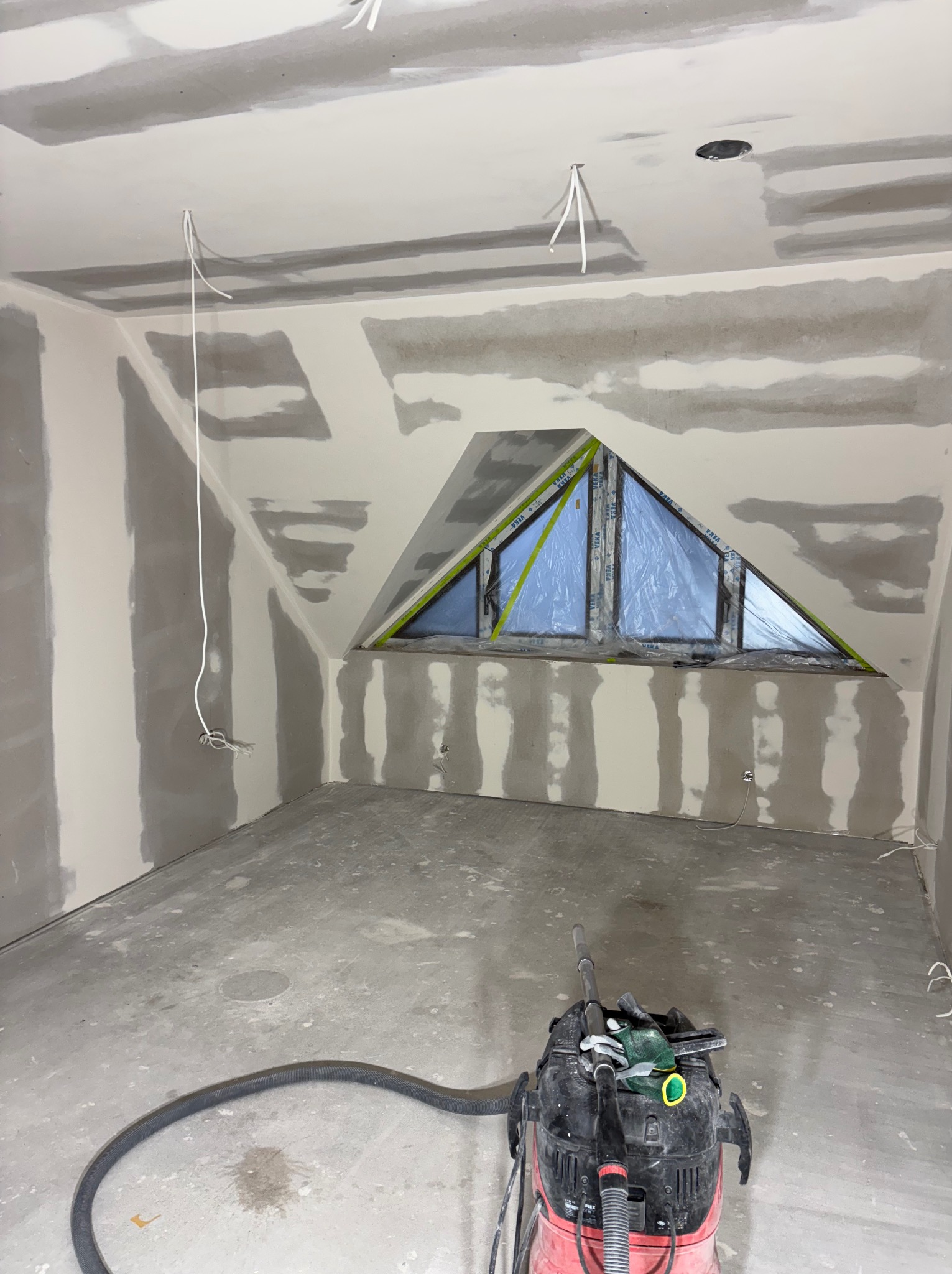 Pokój na poddaszu w trakcie prac wykończeniowych: ściany z płyt gipsowo-kartonowych, szpachlowane łączenia, trójkątne okno zabezpieczone folią, odkurzacz przemysłowy na podłodze.
