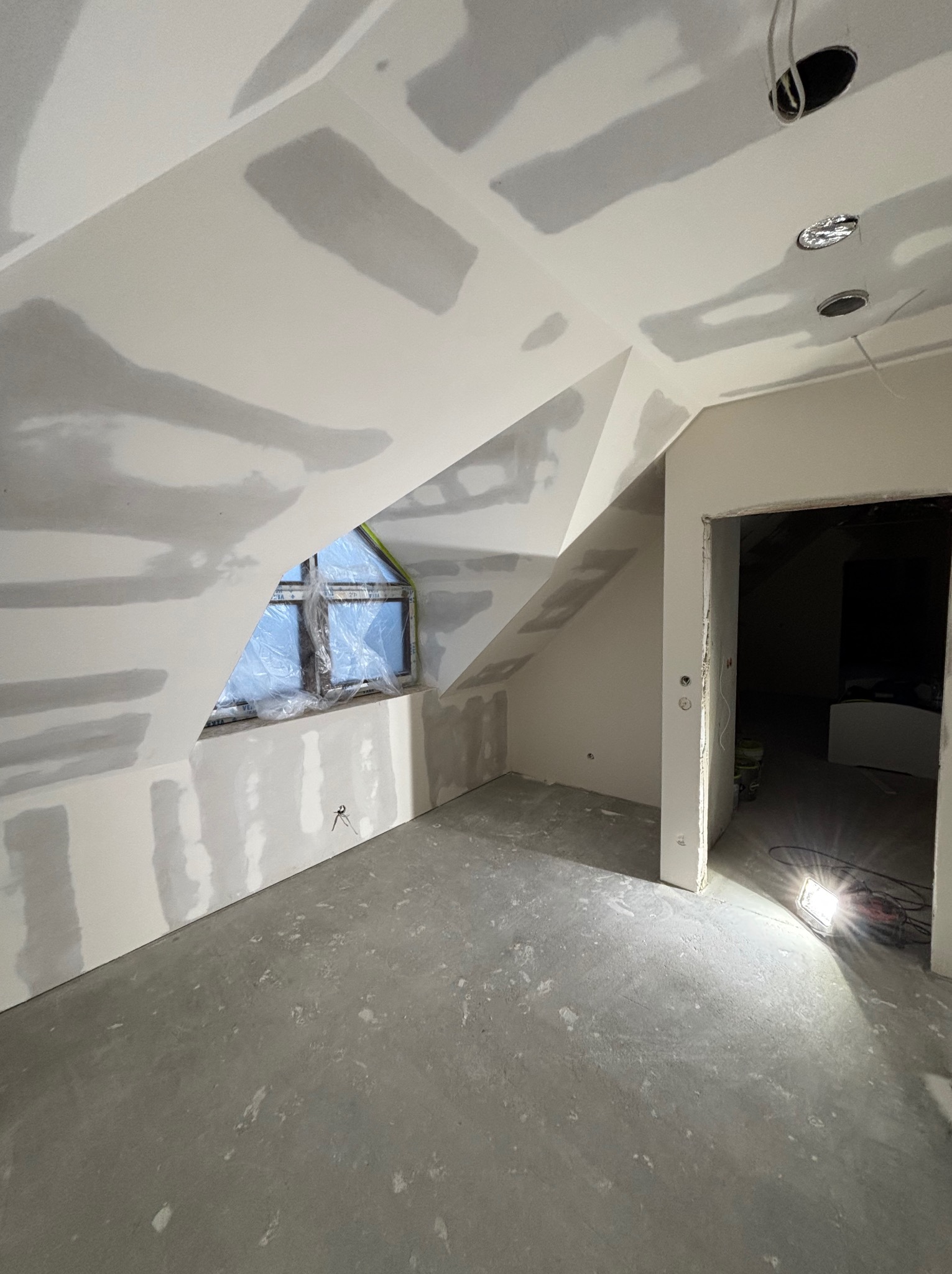 Poddasze w trakcie wykańczania: ściany z płyt gipsowo-kartonowych po szpachlowaniu, okno zabezpieczone folią, surowa wylewka. Widoczne otwory na oświetlenie.