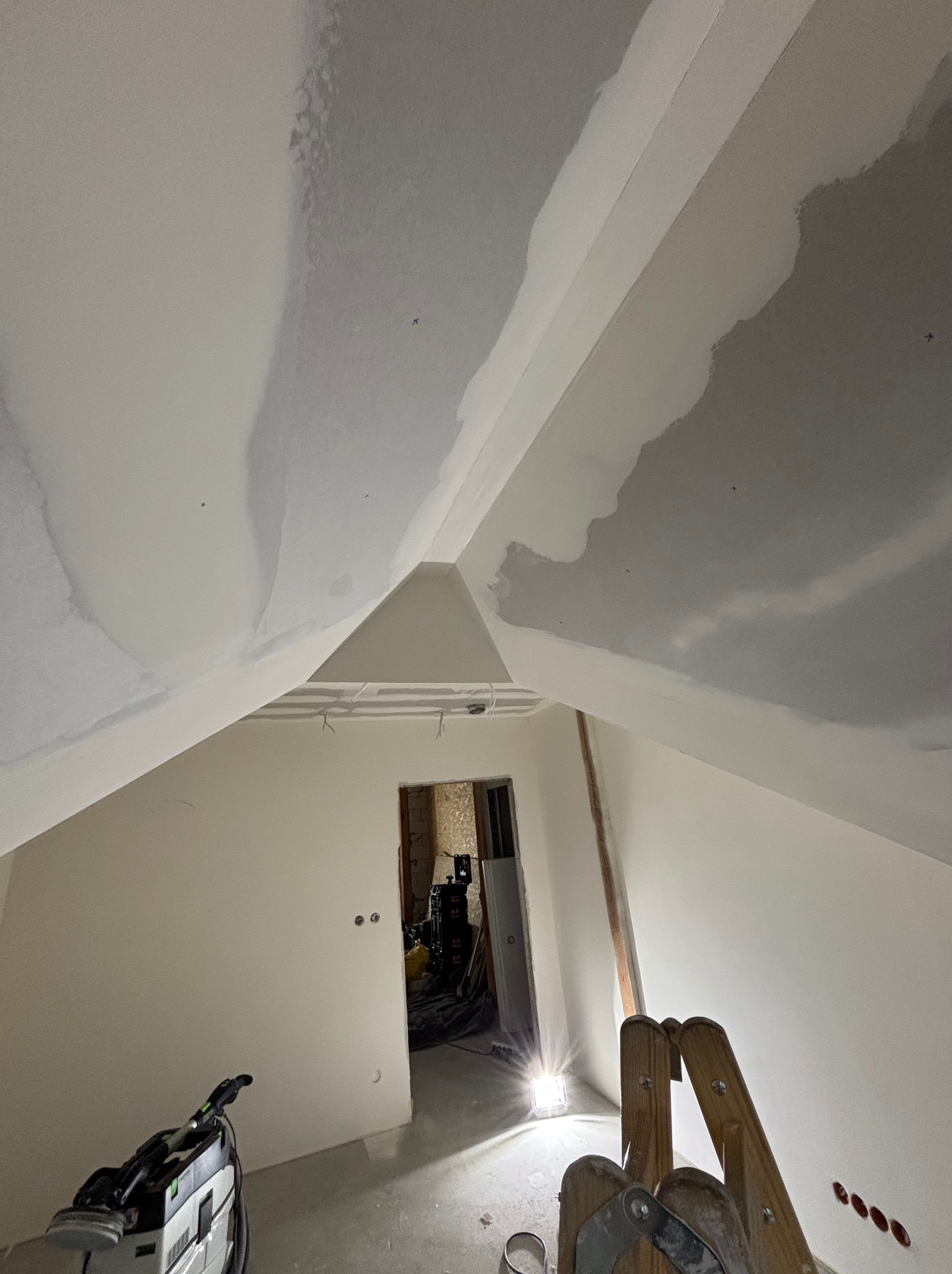 Poddasze w trakcie prac wykończeniowych: szpachlowana skośna ściana i sufit z widocznymi łączeniami płyt, drabina i szlifierka do gładzi na podłodze.