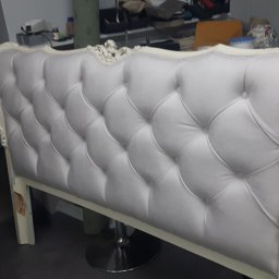 PRACOWNIA TAPICERSKA Wojciech Czapnik - Elegancki zagłówek łóżka z białym, pikowanym obiciem i ozdobną ramą, ustawiony na metalowej podstawie w warsztacie, obok widoczna maszyna do szycia.