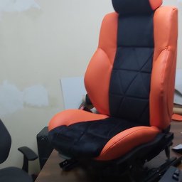 PRACOWNIA TAPICERSKA Wojciech Czapnik - Pomarańczowo-czarne, odnowione siedzenie samochodowe z wyraźnym pikowaniem, widoczne mechanizmy regulacji, w warsztatowym otoczeniu.