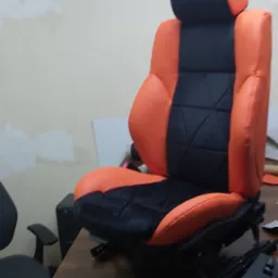 Pomarańczowo-czarne, odnowione siedzenie samochodowe z wyraźnym pikowaniem, widoczne mechanizmy regulacji, w warsztatowym otoczeniu.