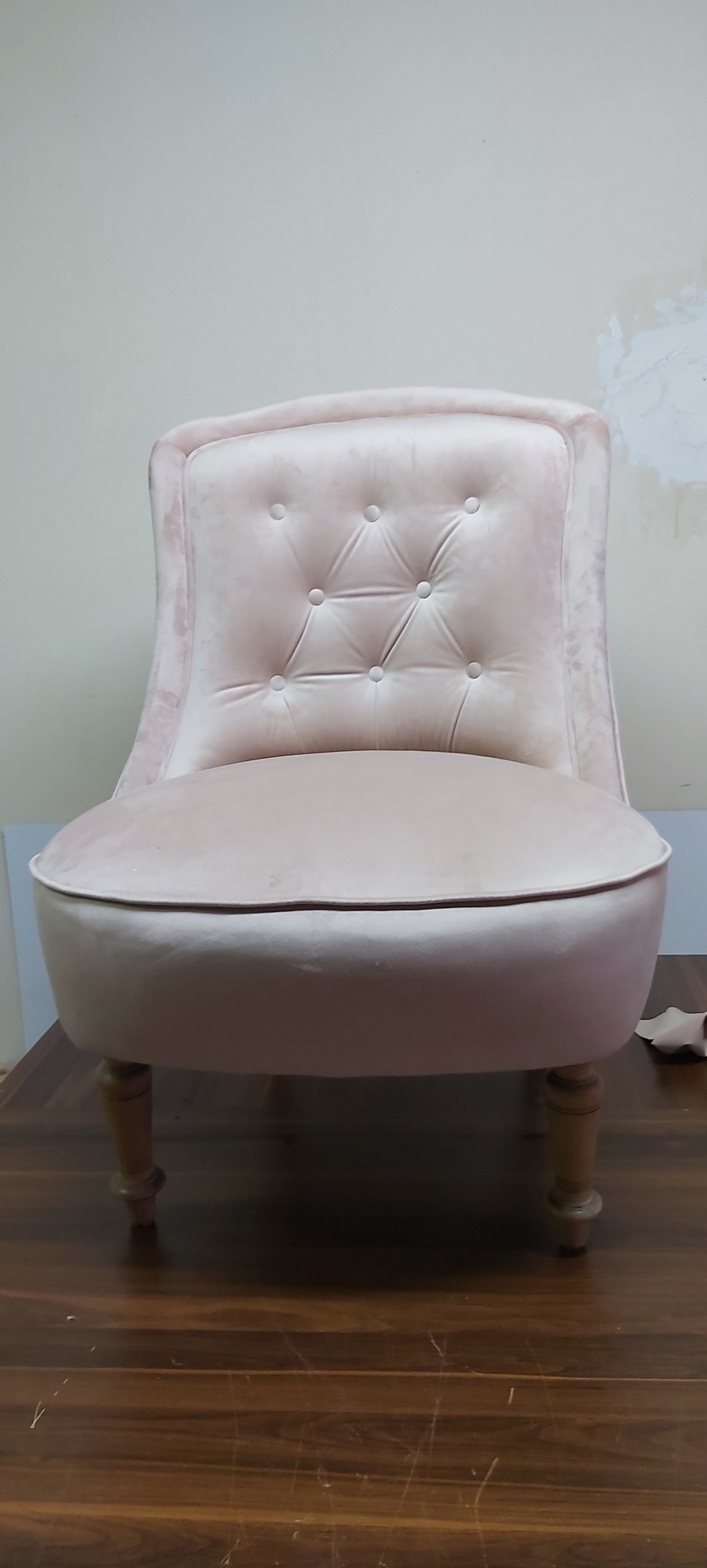 Różowy, tapicerowany fotel w stylu retro z guzikami na oparciu, stojący na brązowej podłodze, na tle jasnej ściany.