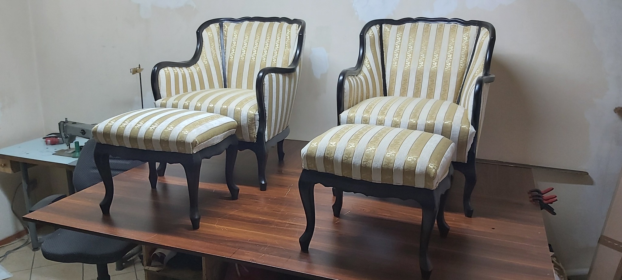 Dwa krzesła i dwa podnóżki z nową, kremowo-złotą tapicerką w paski, stojące na drewnianym stole; w tle maszyna do szycia.