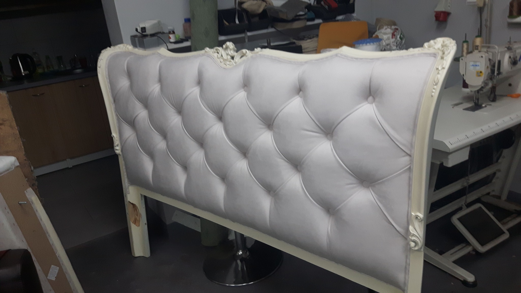 Elegancki zagłówek łóżka z białym, pikowanym obiciem i ozdobną ramą, ustawiony na metalowej podstawie w warsztacie, obok widoczna maszyna do szycia.